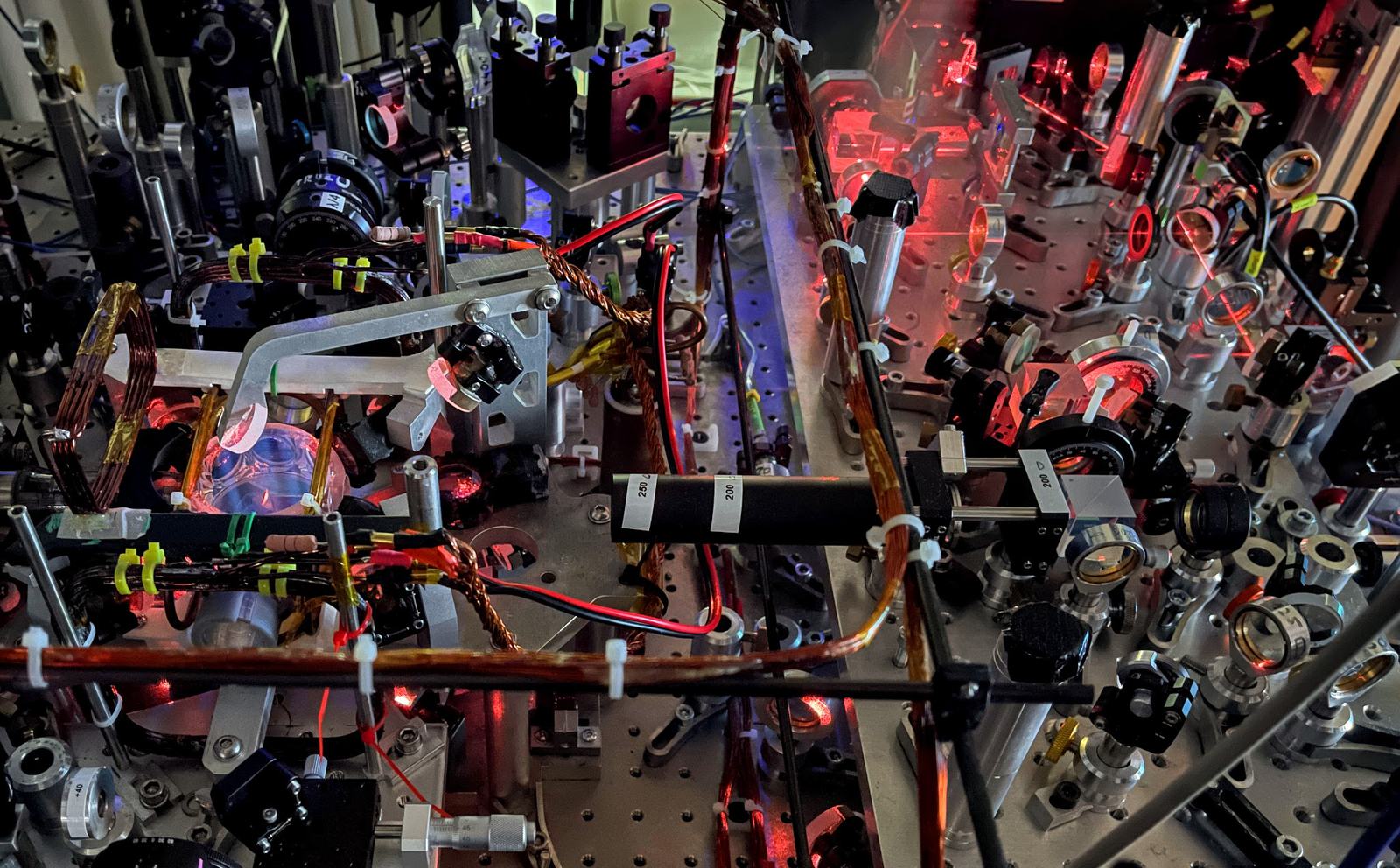 Densely packed optical table with lenses, mirrors and cables, lit by red laser light