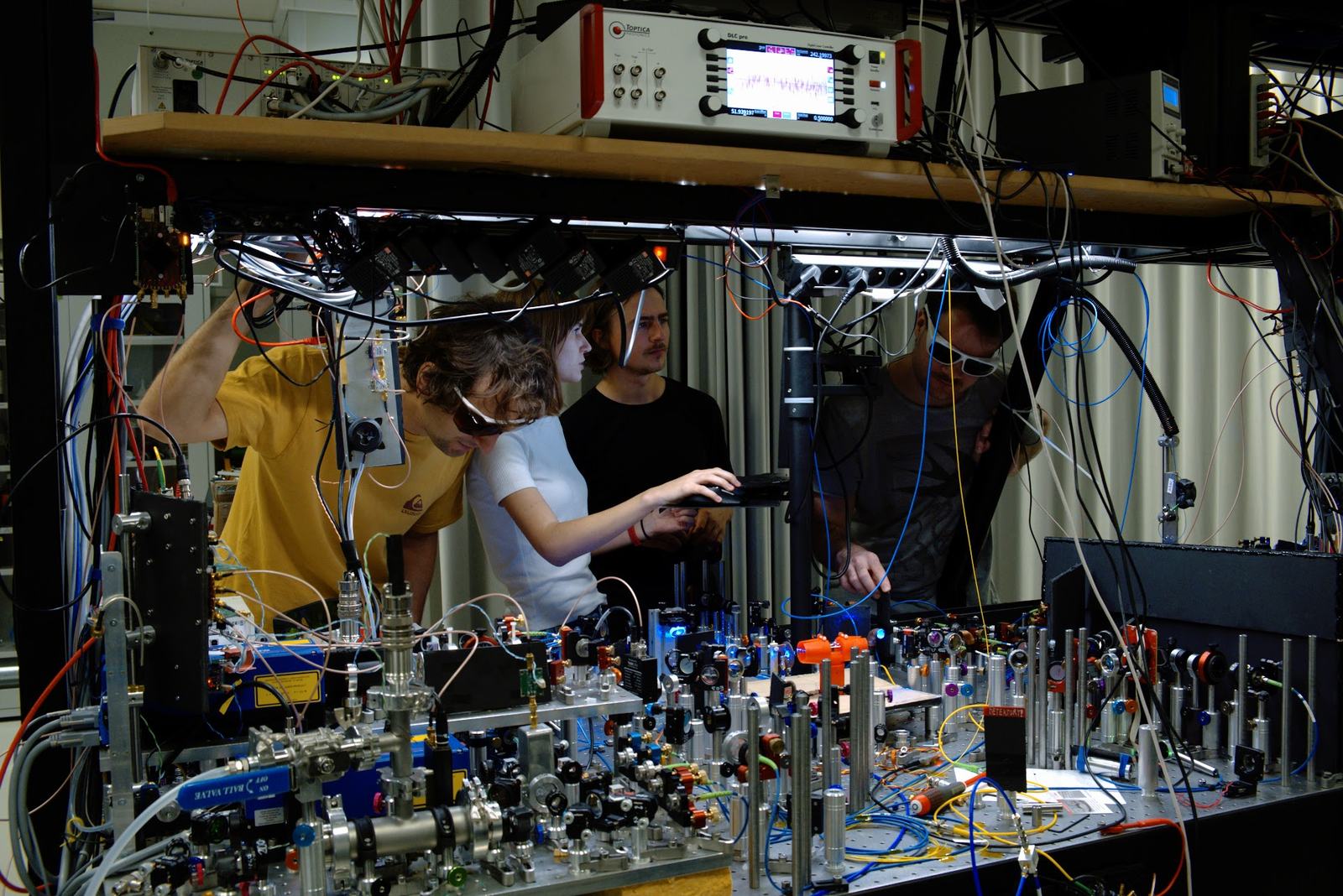 Four researchers working at an optical table under a shelf of electronics in the lab